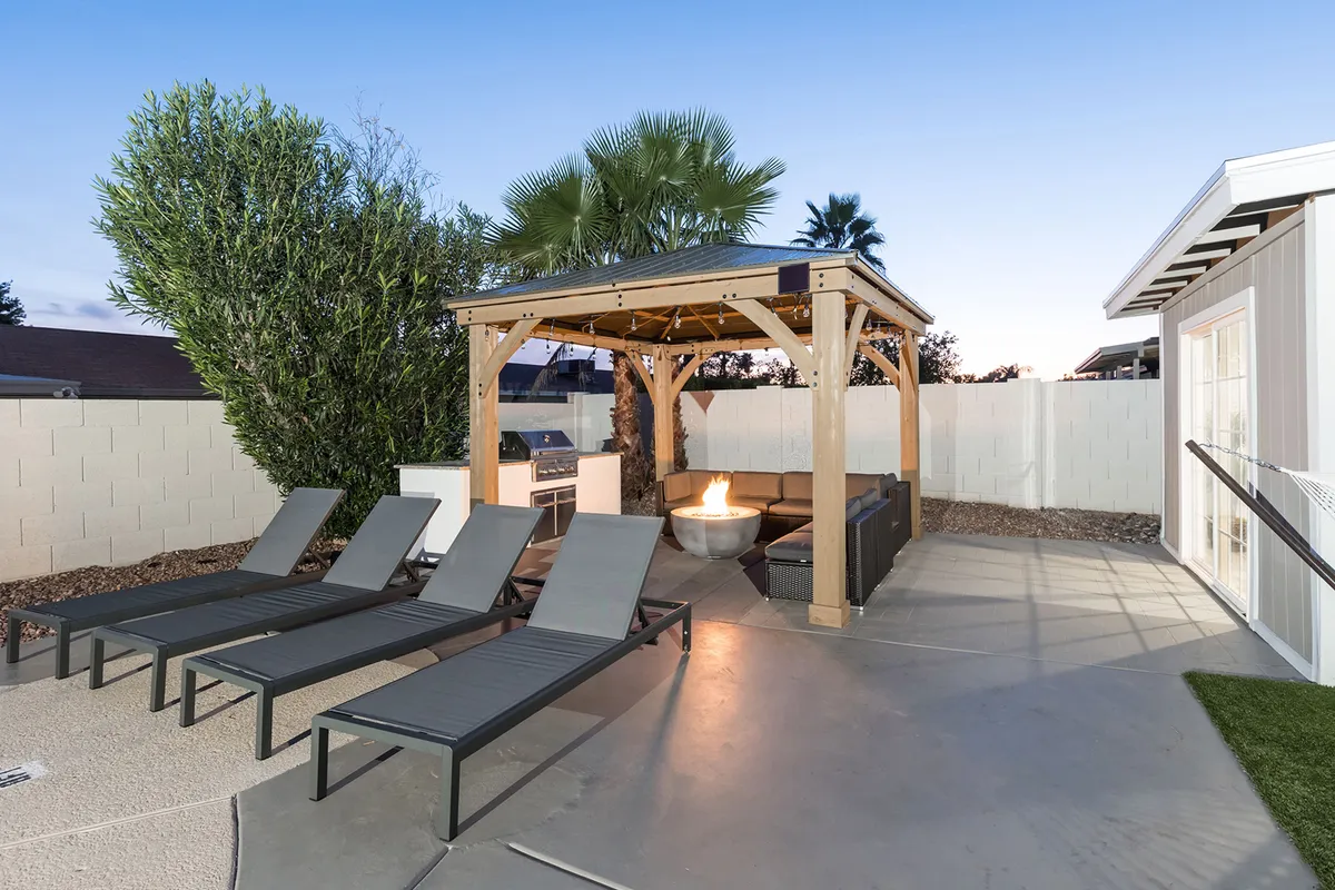 An elegant outdoor seating area under a pergola, featuring a modern fire pit, comfortable lounge chairs, and lush palm trees. The ambient lighting suggests an inviting atmosphere for evening gatherings or intimate conversations under the stars.