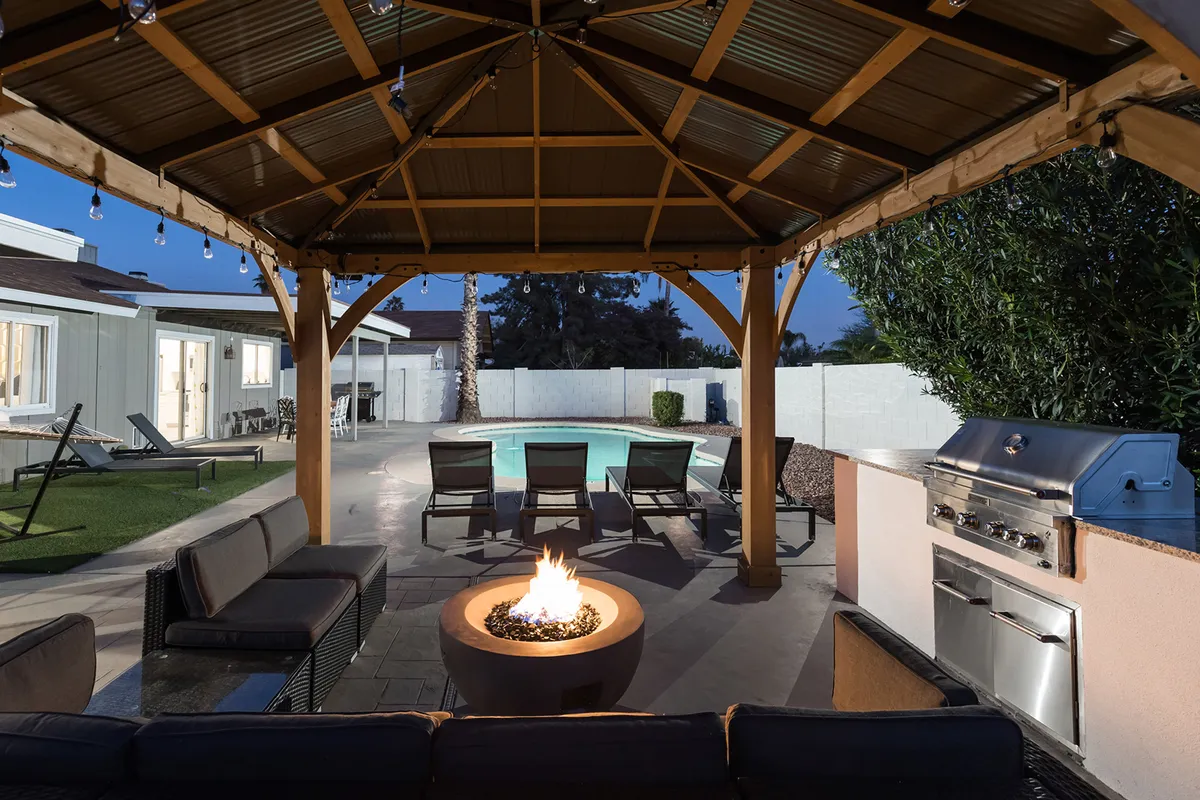 An inviting backyard scene featuring a glowing fire pit surrounded by ample outdoor seating, a shimmering swimming pool, and a grilling station set under the open sky. The area is designed for group gatherings with cozy chairs arranged for conversation and celebration.