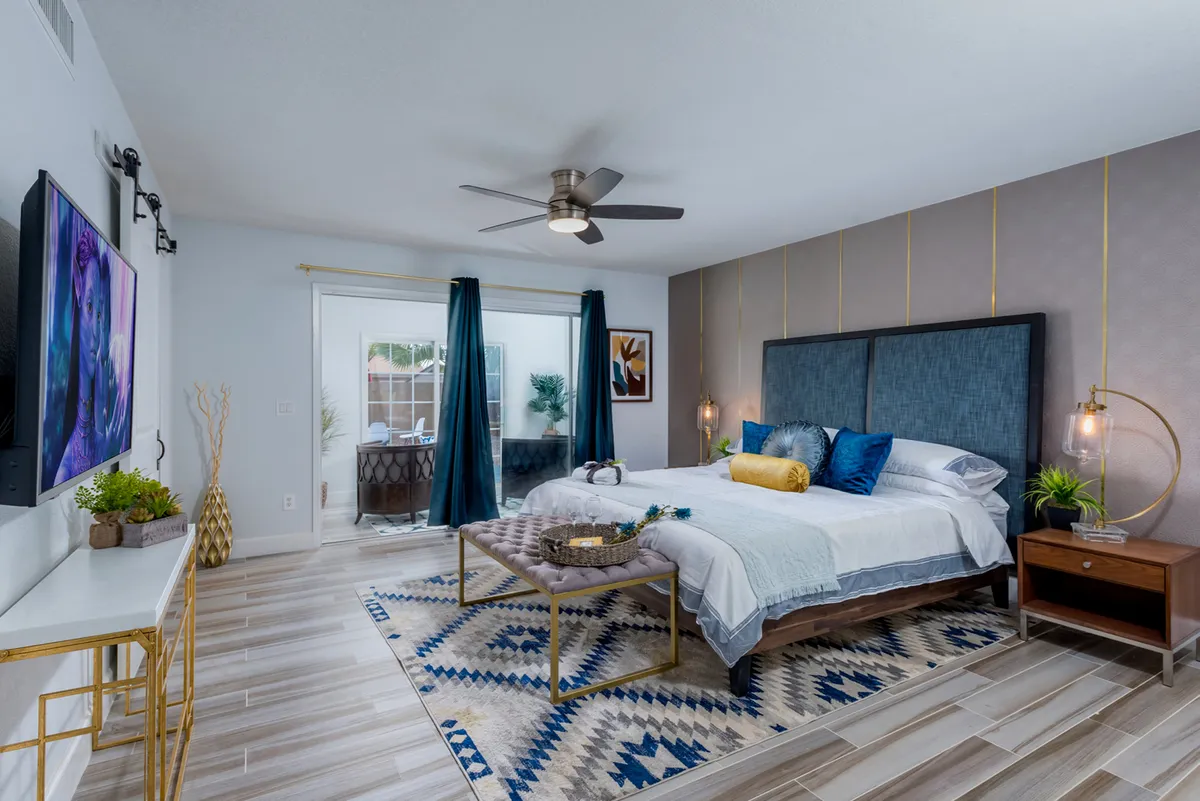 A serene bedroom featuring a large bed, ceiling fan, soft rug, and a television. Sliding doors let in natural light, while the calming decor creates a tranquil atmosphere ideal for relaxation and restful sleep.