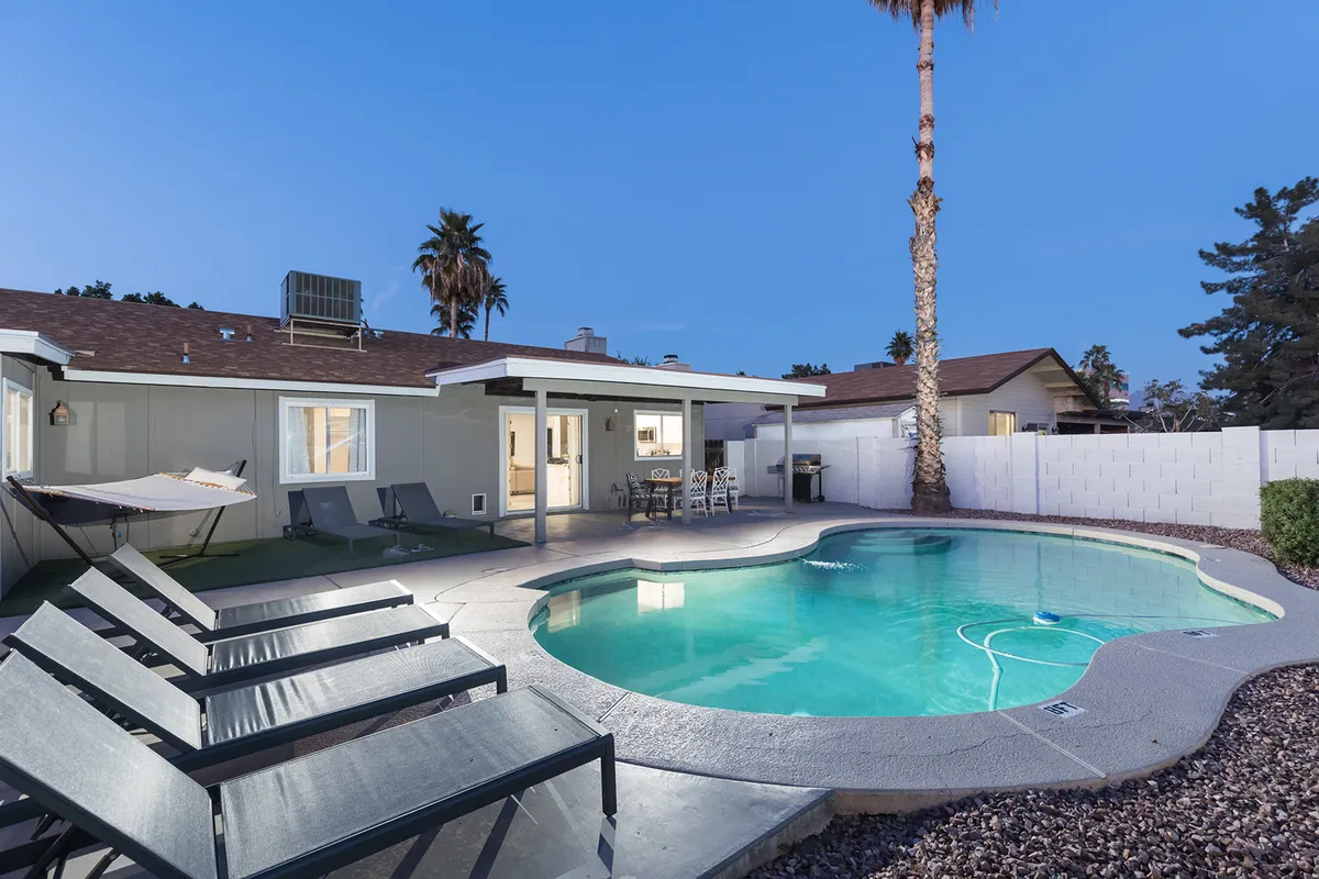 A sun-drenched backyard with a shimmering swimming pool, hammock, lounge chairs, and a spacious patio area surrounded by palm trees. The setting invites relaxation, outdoor socializing, and tranquil poolside moments, with hints of sunset lighting enhancing the vacation vibe.