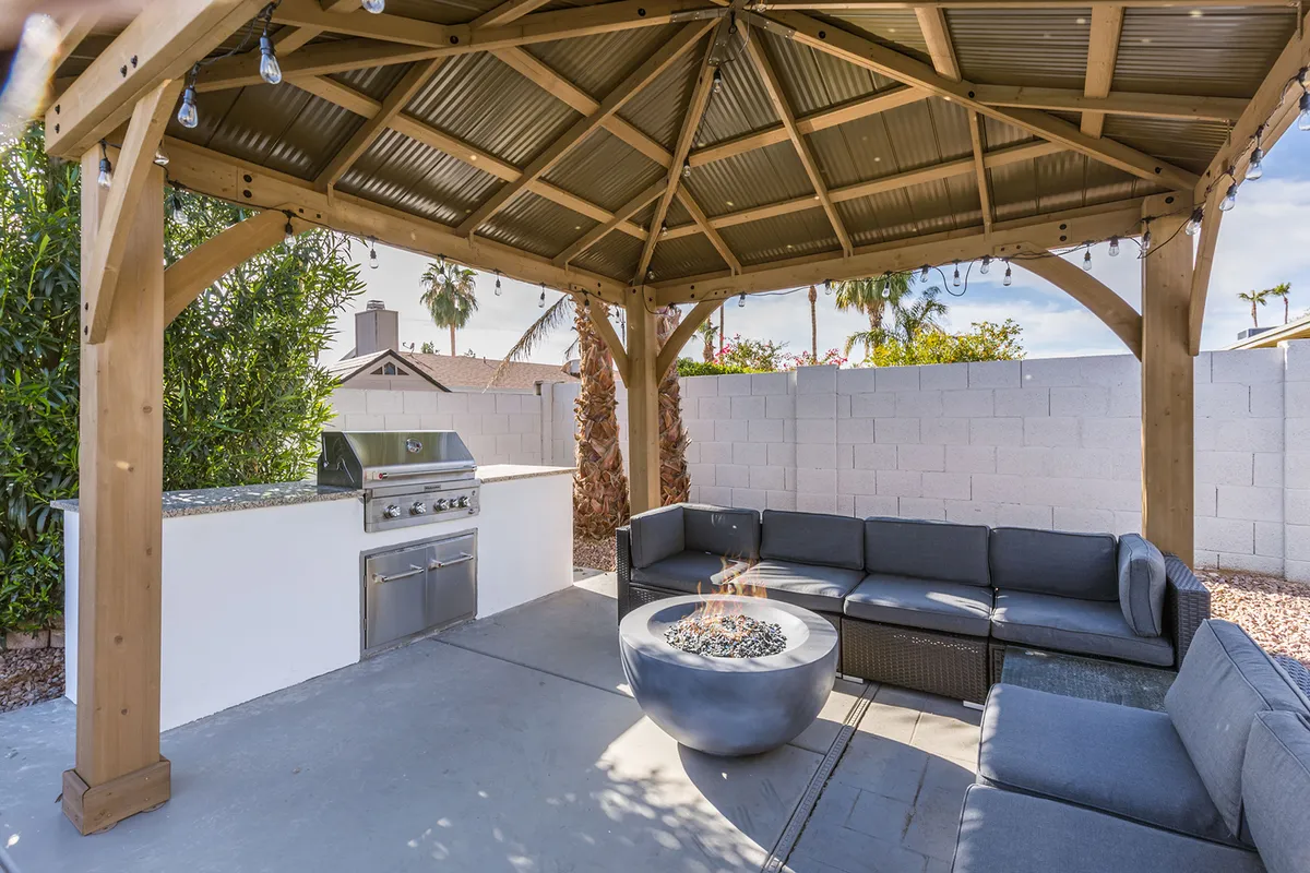 Featuring a wooden gazebo structure with a comfortable outdoor seating area, barbecue grill, and a fire pit, this image spotlights the backyard’s social and entertainment zones. The arrangement is warm and welcoming, ideal for cookouts, celebrations, or simply enjoying the outdoors in comfort.
