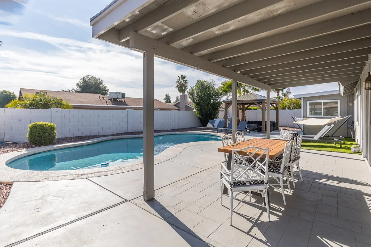 A vibrant backyard with a sparkling swimming pool, an outdoor dining table under a covered patio, and a comfortable lounge area. Palm trees and lush landscaping create a resort-like atmosphere perfect for families to gather, swim, and dine outdoors.